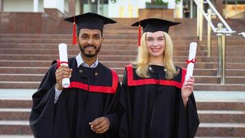 Portrait of happy multiracial graduates. Two friends in graduation caps and gowns standing outside university building, holding diploma scrolls, and smiling video