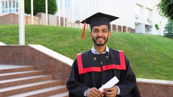 Smiling graduate indian student in graduations gown and hat walking with diploma in campus video