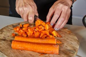 Person chopping fresh carrots on a wooden cutting board in a kitchen setting, with a red peeler nearby. photo