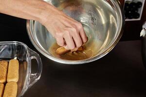 A woman prepares tiramisu dessert in a glass bowl with a mixing bowl on the side in a home kitchen. photo