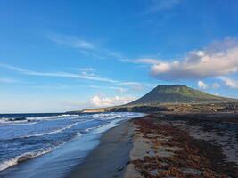 A serene beach with waves lapping at the shore, framed by a lush mountain against a clear blue sky. photo