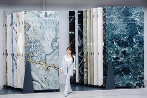 A woman in a light blue suit stands in front of a display of marble tiles. The tiles are arranged in rows and columns, showing a variety of colors and patterns photo