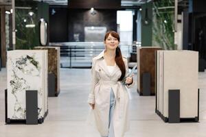 A woman in a white trench coat walks through a showroom displaying tile samples. She is holding a folder in her left hand and smiling at the camera photo