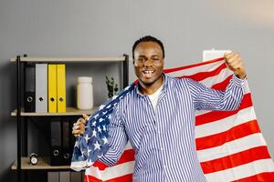 A smiling black man stands in office with an american flag backdrop in hands photo
