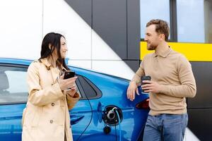 Young couple man and woman traveling by electric car having stop at charging station. Boyfriend plugging in cable to charge. Man talking with girlfriend, holding cup drinking hot coffee smiling photo