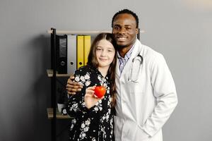 Man in white robe and girl standing together, girl holding red heart photo