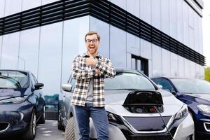A young man stands next to his new electric car and rests while the car is charging photo