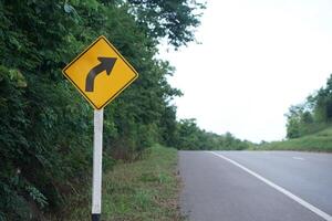 Yellow traffic sign with arrow left curve symbol beside the rural road to warn drivers to know the way ahead has right curve. Concept. Warning traffic sign for transportation. photo