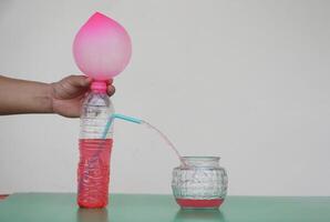 Science experiment with air in pink balloon on the top of bottle with red water and straw tube to put water flow into glass jar. Concept, teaching aids, educational materials. Creative science lesson. photo