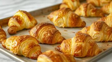 Golden flaky croissants fresh out of the oven are placed on a lined baking sheet. The pastries have a perfect crispy exterior, showcasing their multiple layers and buttery texture, ideal photo