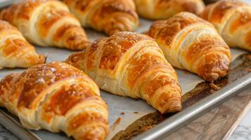 Golden flaky croissants fresh out of the oven are placed on a lined baking sheet. The pastries have a perfect crispy exterior, showcasing their multiple layers and buttery texture, ideal photo