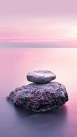 A single stone rests on a larger rock, overlooking a calm body of water during sunset photo