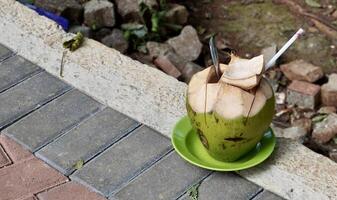 Kelapa muda juice water and fruit snack with green plate, straw, and stainless spoon isolated on horizontal ratio brick side walk ground flooring background. photo