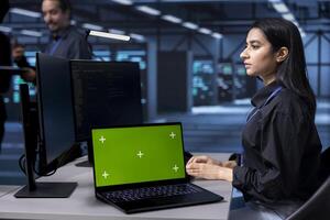 Admin working on green screen laptop in data center housing storage devices. Woman using chroma key notebook in facility with networking components providing vast amounts of computing resources photo