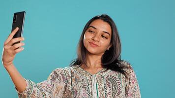 Cheerful indian woman using smartphone to take selfies and post them on social media. Happy person taking photos using phone selfie camera, isolated over studio background, camera B