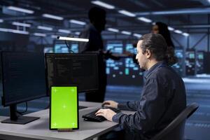 Programmer works on green screen tablet in data center housing storage equipment. IT professional using mockup device in server farm with networking components providing vast amounts of computing resources photo