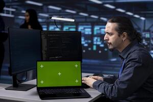 Engineer working on isolated screen laptop in data center housing storage devices. Man using mockup notebook in facility with networking components providing vast amounts of computing resources photo
