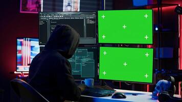 Hacker developing spyware software on green screen computer to steal important data to be sold on black market. Hooded scammer writing malicious code on chroma key PC, camera A photo