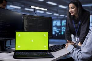 Focus on green screen notebook used by engineers in blurry background to oversee servers used for managing databases. Close up shot of mockup laptop used by IT specialists in data center photo