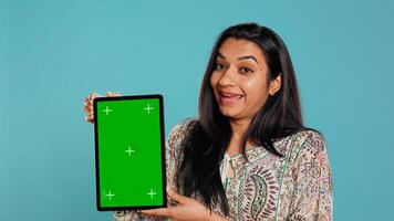 Portrait of indian woman doing influencer marketing using green screen tablet, isolated over studio background. Smiling person holding empty copy space mockup device, camera B photo