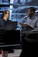 Manager getting feedback from engineer while overseeing data center, looking for firewall misconfigurations. Supervisor talking with workers in server room, asking about systems bottlenecks photo