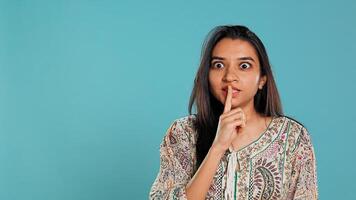 Annoyed indian woman doing shushing hand gesturing, irritated by noise, having negative mood. Person placing finger on lips, doing quiet sign gesture, isolated over studio background, camera B photo