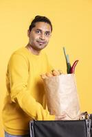 Portrait of cheerful deliveryman taking takeaway order out of bag, providing it to client. Joyous person taking groceries order out of thermal backpack, ordered by customer, studio backdrop photo