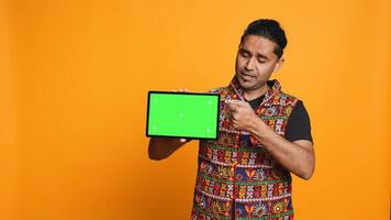 Portrait of indian man doing influencer marketing using green screen tablet, isolated over studio background. Smiling person holding empty copy space mockup device, camera B photo