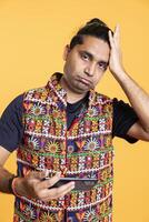 Gamer playing online multiplayer videogame on smartphone, annoyed after losing, being defeated by foes. Indian person stumped after seeing game over message, holding mobile phone, studio background photo