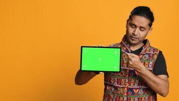 Portrait of indian man doing influencer marketing using green screen tablet, isolated over studio background. Smiling person holding empty copy space mockup device, camera A photo