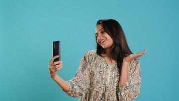 Jolly indian woman smiling for selfies, posting them on social media using cellphone. Joyous girl taking pictures using phone selfie camera, isolated over studio background, camera A photo