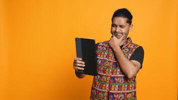Relaxed man having entertaining conversation with friend over online videocall on device. Carefree indian person discussing with mate on teleconference using tablet, studio backdrop, camera B photo