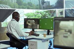 African american operator monitoring traffic through CCTV surveillance next to a greenscreen display, security cameras footage at stop lights. Employee gathering data on cars via satellite. photo