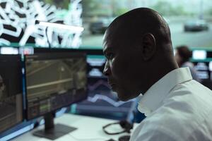 African american employee monitoring traffic and locating truck driver, updating clients after tracking order in control center. Male operator uses CCTV surveillance footage, gps tracing. photo