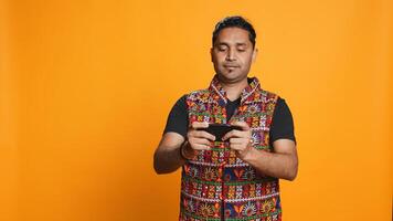 Indian man playing videogames on cellphone in landscape mode, enjoying leisure time. Gamer enjoying game on mobile phone, having fun, isolated over studio background, camera B photo