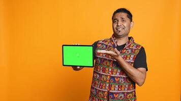 Upset indian man shows thumbs down sign gesturing holding isolated screen tablet, disapproving. Person does rejection hand gesture regarding mockup device, studio background, camera A photo