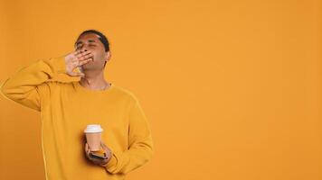 Indian man enjoying coffee from disposable paper cup and send text messages on smartphone. Person drinking hot beverage and chatting with friends on mobile phone, studio background, camera A photo
