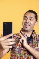 Joyous indian narcissistic man using cellphone to take selfies from all angles. Vain social media user taking photos using phone selfie camera, smiling happily, studio background