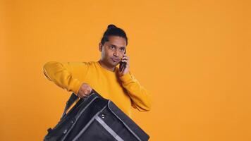 Man packing food in thermal backpack, fulfilling clients orders and answering phone ringing, studio backdrop. Indian person doing delivery to customers, discussing with them on cellphone, camera B photo