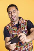 Man in traditional clothing entertained by videogames on smartphone, delighted by action. Player sticking tongue out while enjoying game on phone, having fun online, studio backdrop photo