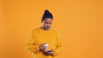 Indian man enjoying coffee from disposable paper cup and send text messages on smartphone. Person drinking hot beverage and chatting with friends on mobile phone, studio background, camera B photo