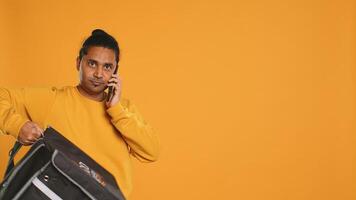 Man packing food in thermal backpack, fulfilling clients orders and answering phone ringing, studio backdrop. Indian person doing delivery to customers, discussing with them on cellphone, camera A photo