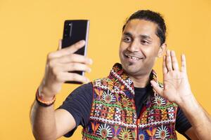 Joyous man doing waving hand gesture, talking with friend over internet videocall on smartphone. Upbeat indian person saluting mate during videoconference using phone, studio background photo
