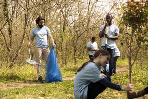 Environmental volunteers collecting rubbish and plastic waste in garbage bag, using littler cleanup tools to pick up trash from the woods. Diverse people doing voluntary work to protect ecosystem. photo