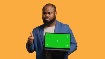 Salesman holding isolated screen laptop, doing recommendation, talking with audience. BIPOC person pointing towards chroma key notebook and showing thumbs up sign, studio background, camera A photo