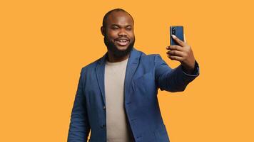 Joyous man using smartphone to take selfies and post them on social media. Happy BIPOC person taking photos using phone selfie camera, isolated over yellow studio background, camera A