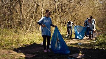 Small girl collecting rubbish around a forest with a claw tool and garbage bags, cleaning up the environment by recycling plastic waste and trash. Child learning to protect ecosystem. Camera B. photo