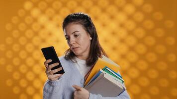 Smiling woman reading messages on smartphone while holding stack of books, isolated over studio background. Cheerful person with pile of academic textbooks in arms texting friends on phone, camera B photo
