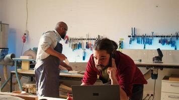 Cabinetmaker receiving telephone call from client inquiring about furniture assembling commission progress. Carpenter in assembly shop talking on smartphone with customer, camera B photo