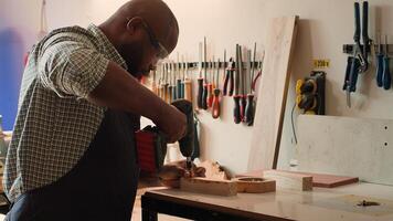African american man in carpentry shop using power drill to create holes for dowels in wooden board. Craftsperson sinks screws into wood block with electric tool, doing precise drilling, camera A photo